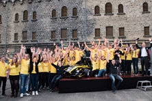 Eine Gruppe von rund 50 Studierenden in einheitlichen, gelben T_Shirts steht um einen schwarz-gelben Rennwagen und hebt die Hände jubelnd in die Höhe.