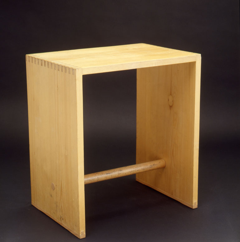 Der Hocker besteht aus zwei parallelen Holzbrettern, die am einen Ende von einem dritten Brett zusammenhalten werden, am anderen Ende von einem dünnen Holzstab.