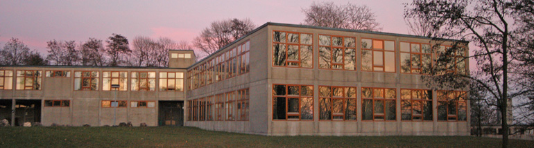 Die grauen Betonquader der Hochschule für Gestaltung mit ihren orange eingerahmten Fenstern im Vordergrund, im Hintergrund lila schattierter Himmel.