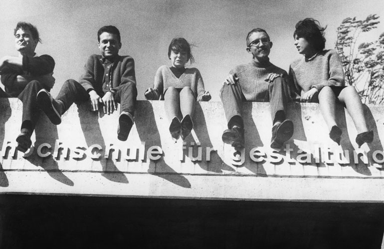 Schwarz-weiß-Foto von fünf Leuten, die auf einer Brüstung mit der Aufschrift "Hochschule für Gestaltung" sitzen. Sie wirken entspannt und gut drauf und scheinen zwischen 20 und 40 Jahren zu sein.